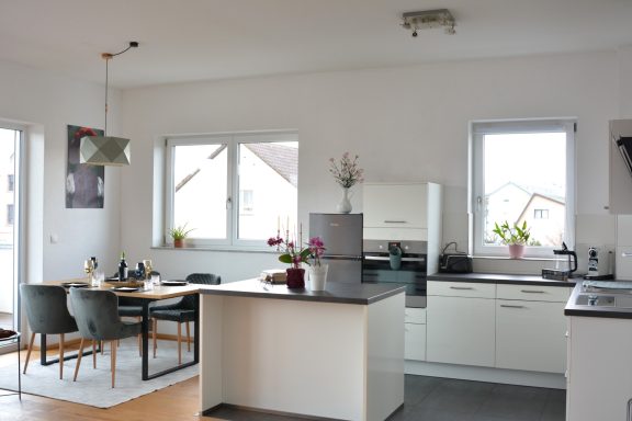 Moderne, helle Küche mit Tisch und Stühlen, großen Fenstern und Pflanzen.