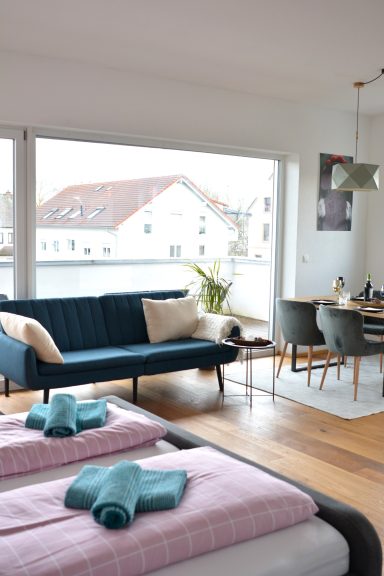Helles, modernes Zimmer mit zwei Betten, Sofa, Esstisch und großen Fenstern.